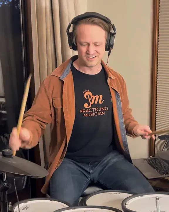 Jake Douglass smiling as he plays an electronic drum kit at home, in a Practicing Musician T-shirt.